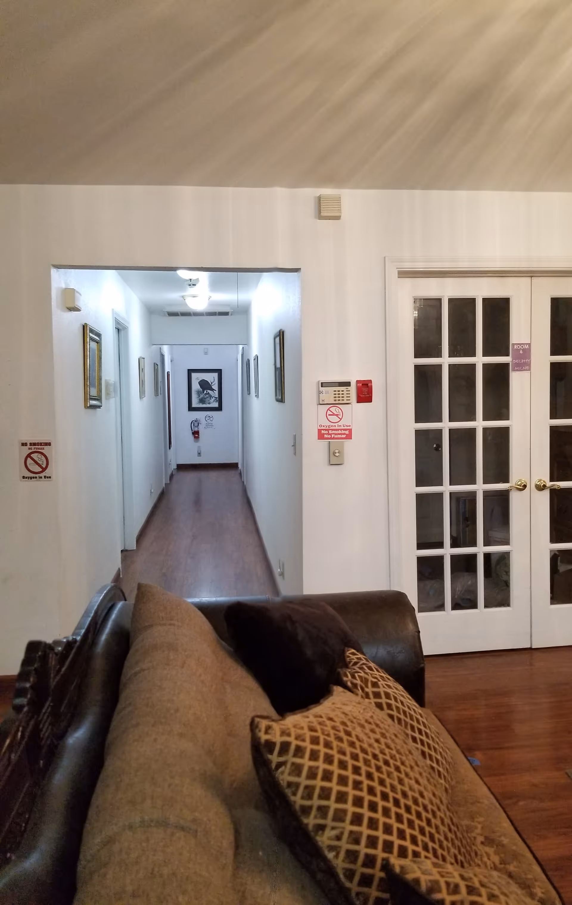 Interior view of a senior living facility hallway with wooden flooring, white walls, and framed pictures on the walls. In the foreground, there is a brown couch with patterned cushions. To the right, there are white double doors with glass panels and a small sign that reads 'ROOM 6 DELPHY MILNE'. There are also safety signs indicating no smoking and oxygen in use.
