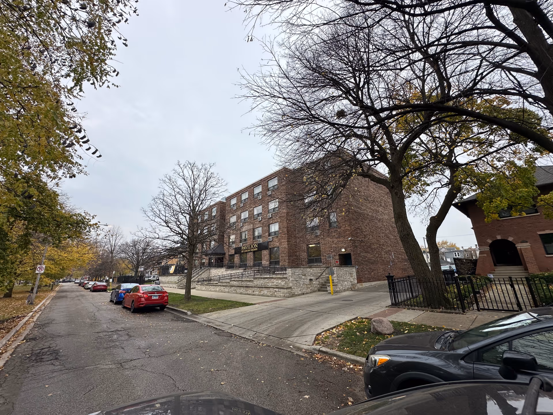 Exterior view of a multi-story brick building named Pavilion of Logan Square, situated on a street lined with parked cars and trees with autumn foliage. The sky is overcast and the building has a stone base with a ramp leading to the entrance.