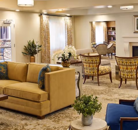 A cozy and well-lit living room area in an assisted living facility featuring a mustard yellow sofa with blue patterned pillows, two patterned armchairs, a blue cushioned chair, a round table with a green plant, and a side table with a white floral arrangement. The room has beige walls, large windows with curtains, and a carpeted floor with a subtle pattern. In the background, there is a fireplace and a small seating area with bookshelves.
