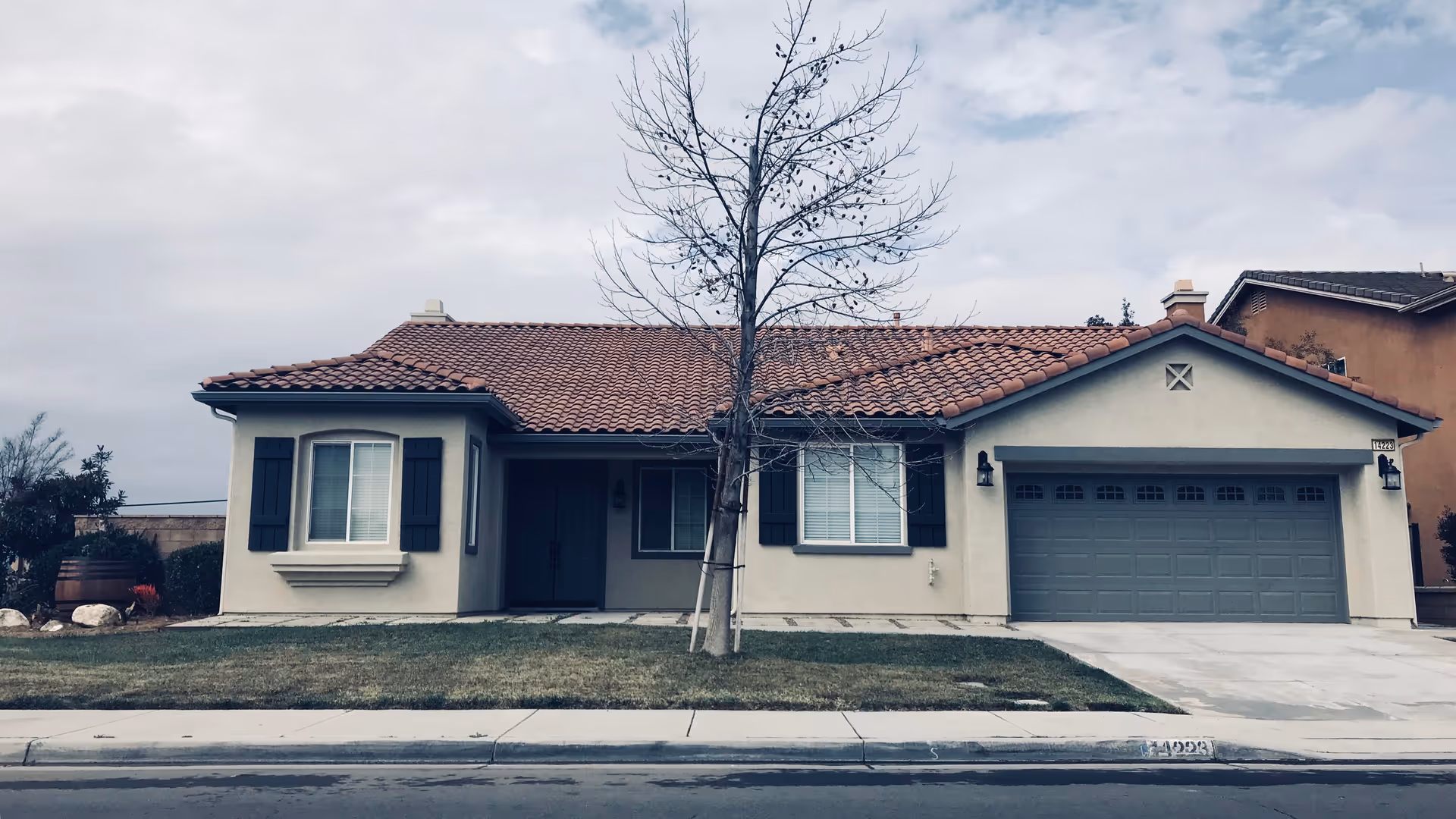 Front exterior view of a single-story house with a tiled roof, two windows with black shutters, a double front door, a garage with a closed door, and a leafless tree in the front yard.