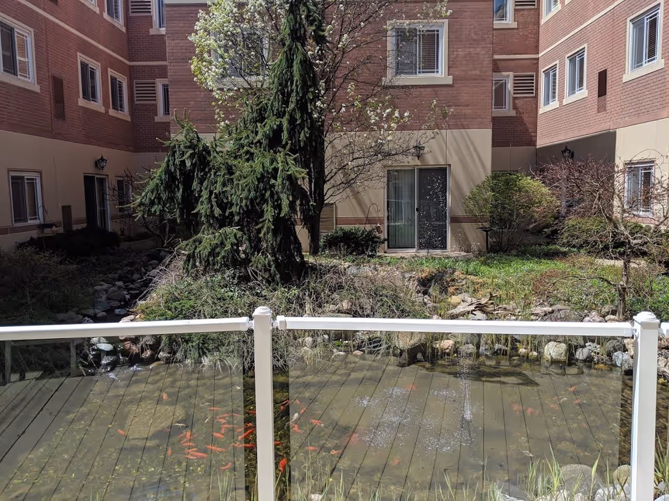 View of an outdoor courtyard at Franciscan Court Supportive Living featuring a small pond with orange fish, a white railing in the foreground, various trees and shrubs, and a multi-story brick building surrounding the courtyard.