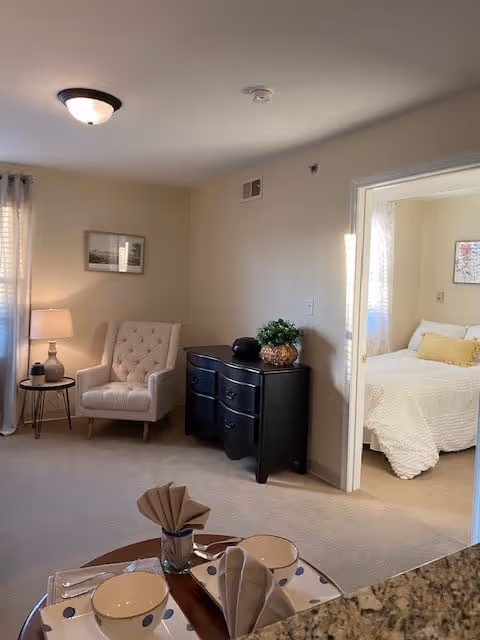 A cozy living area with a beige armchair next to a small round table with a lamp. A black dresser with a plant and decorative items is against the wall. Through an open doorway, a bedroom with a white bedspread and pillows is visible. A dining table with place settings and folded napkins is partially visible in the foreground.