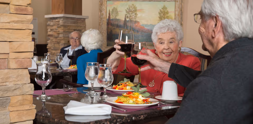 Two elderly people sitting at a dining table in a senior living facility, clinking glasses of red wine. Plates of food and glasses of water are on the table. In the background, two other elderly people are seated at another table. The setting is warm and inviting with stone pillars and a framed landscape painting on the wall.
