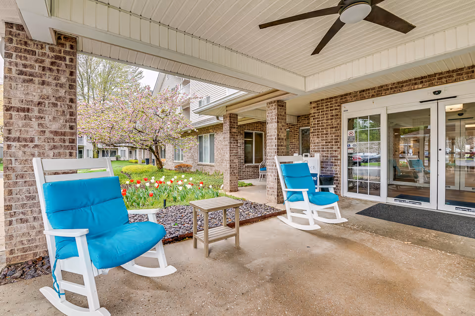 Covered outdoor seating area at the entrance of a senior living facility with two white rocking chairs featuring bright blue cushions, a small wooden table between them, brick pillars, a ceiling fan, and glass double doors leading inside. In the background, there is a garden with blooming tulips and a tree with pink flowers.