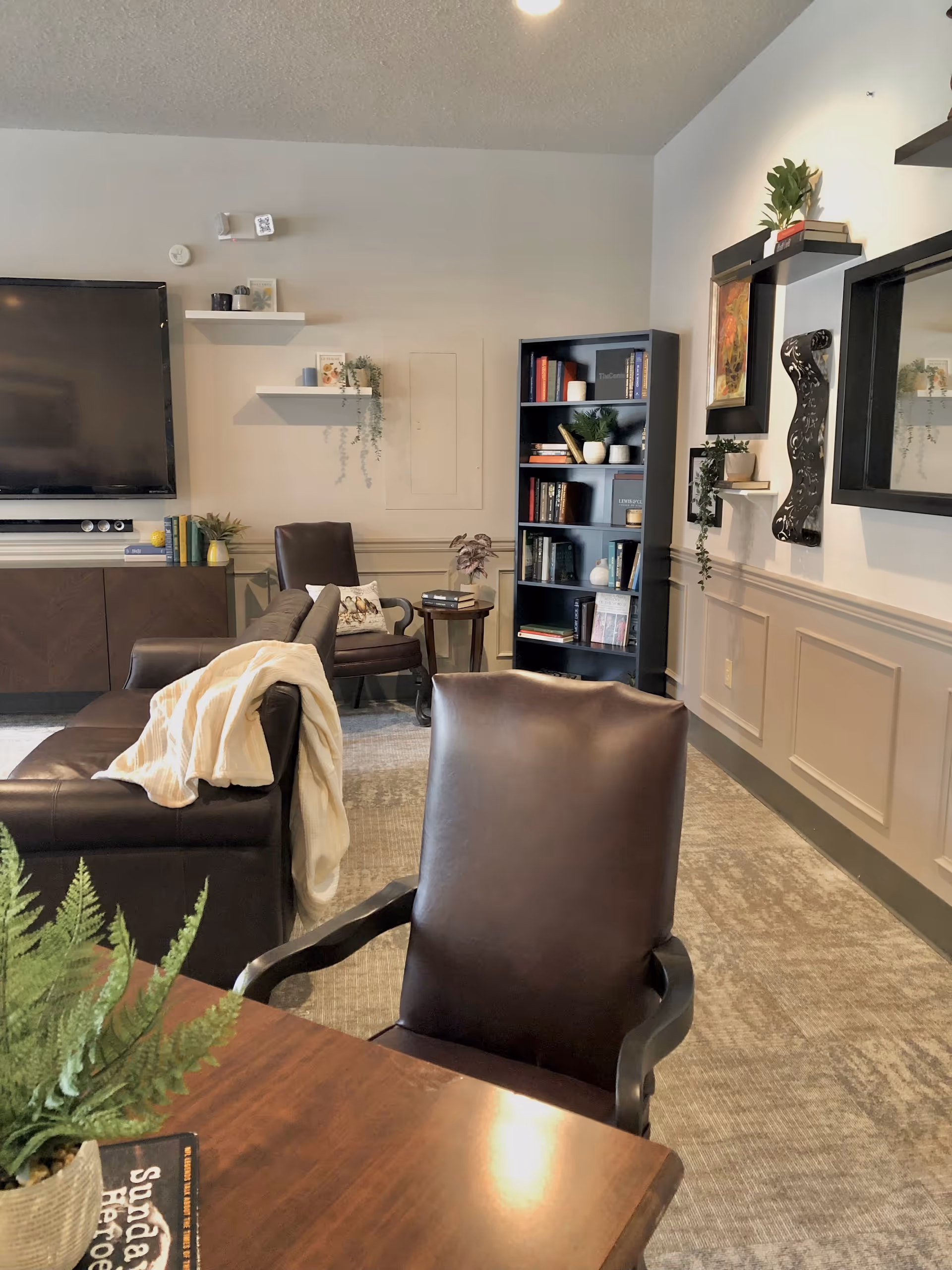 A cozy living room area with a dark leather chair in the foreground, a brown leather sofa with a cream-colored blanket draped over it, a dark wooden table with a potted fern, a large flat-screen TV mounted on the wall, floating shelves with decorative items, a black bookshelf filled with books and plants, and framed artwork on the walls.