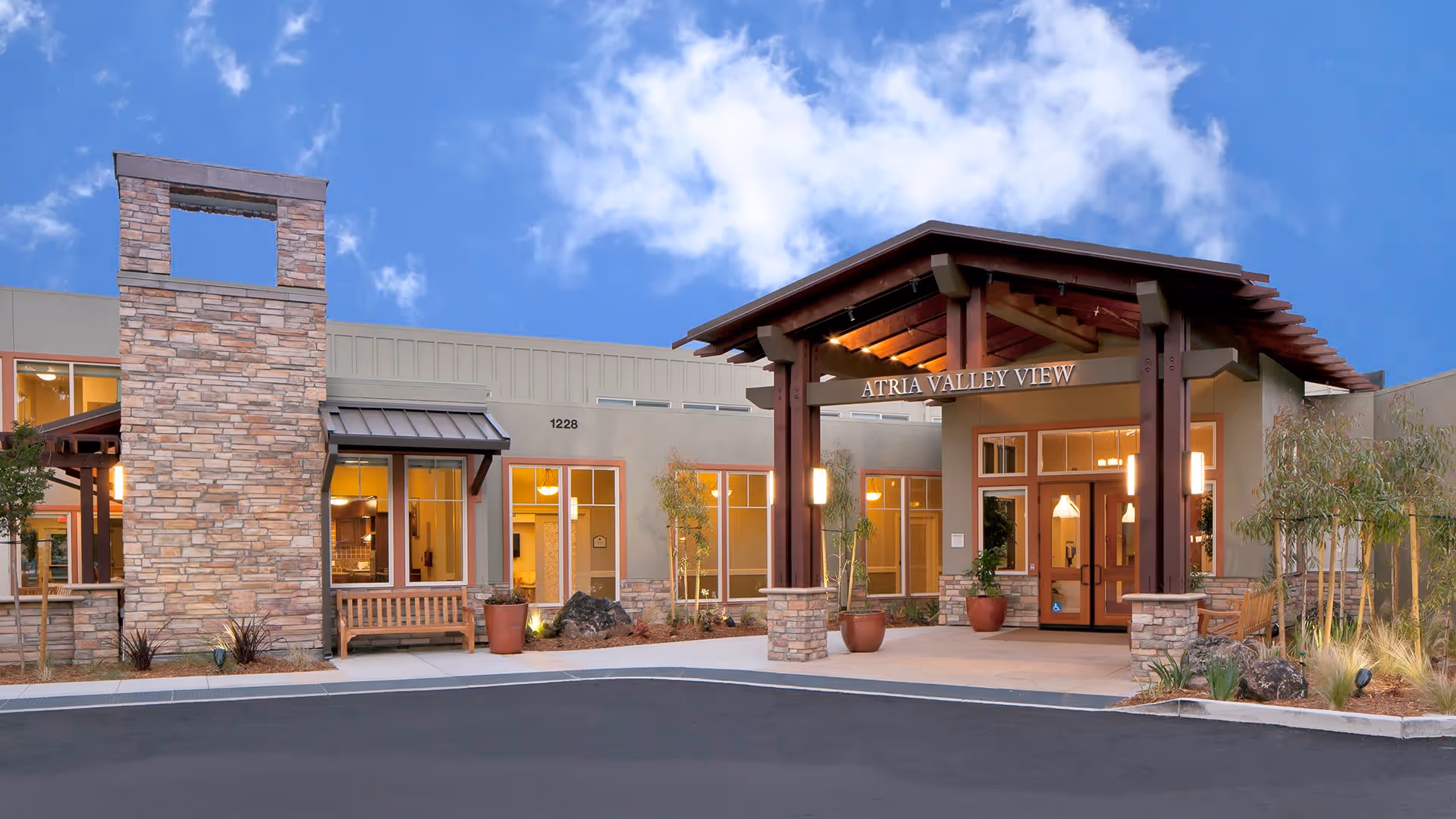 Exterior view of the entrance to Atria Valley View senior living facility, featuring a covered wooden pergola, stone pillars, large windows, benches, and landscaping under a partly cloudy blue sky.