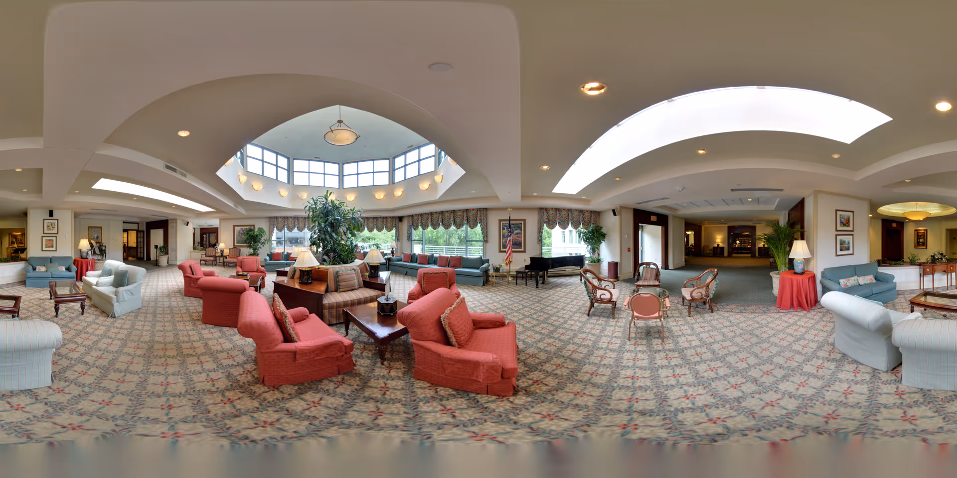 A spacious and well-lit senior living facility common area with a patterned carpet, multiple seating arrangements including red and light blue armchairs and sofas, wooden coffee tables, and lamps. The room features large windows with valances, a skylight, and decorative plants. There is a grand piano near the windows and framed artwork on the walls.