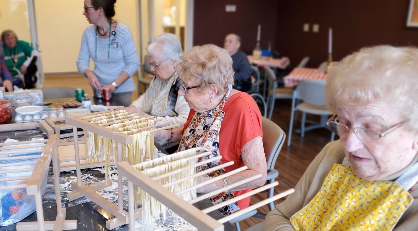 Several elderly women seated around a table in a communal room, engaged in making and drying fresh pasta on wooden racks. A caregiver or staff member stands nearby assisting. The room has tables and chairs with a warm, inviting atmosphere.