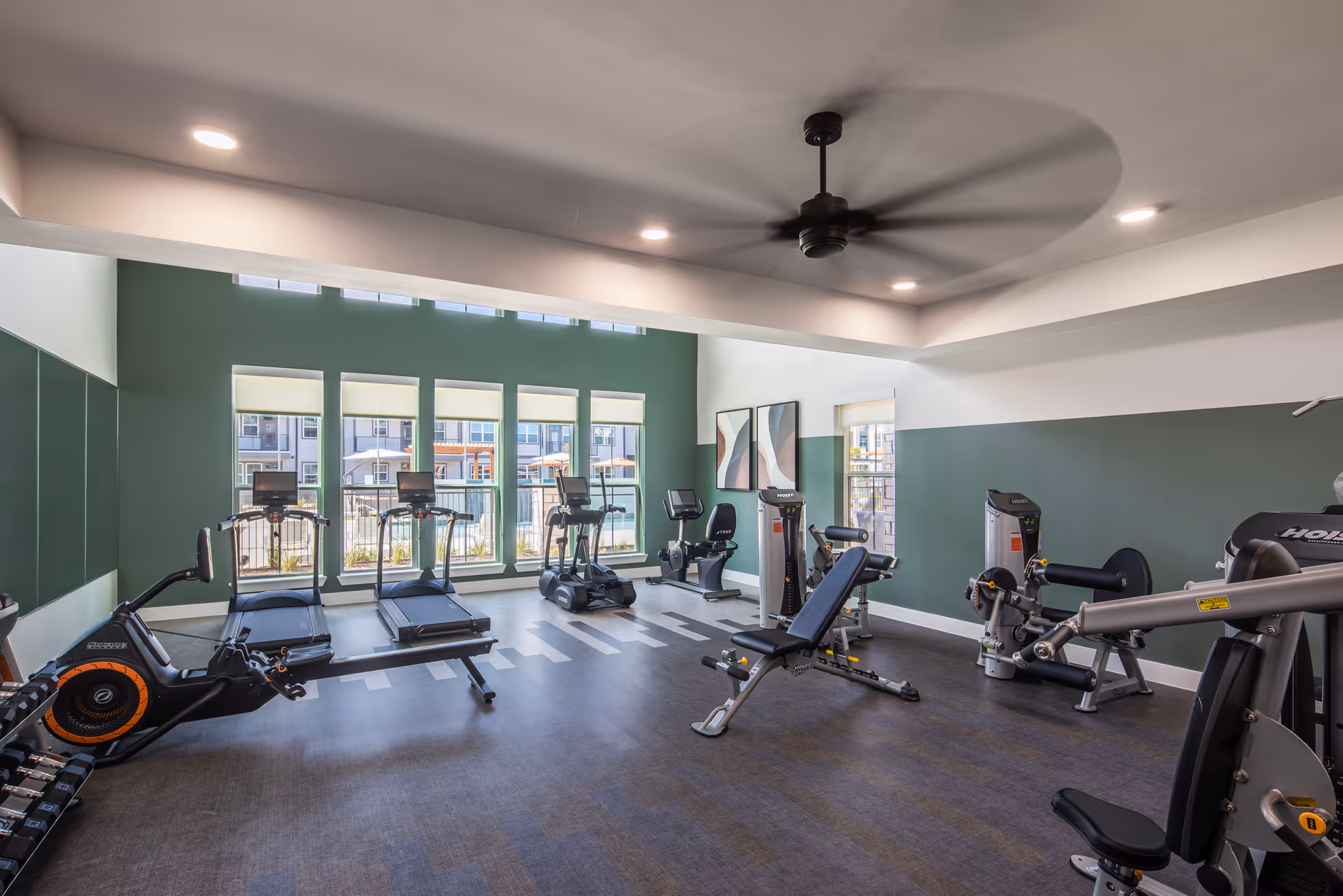 Well-lit fitness room with treadmills, exercise bikes, weight machines, and large windows overlooking the exterior.