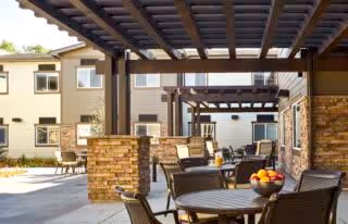 Outdoor patio area at Ralston Creek Neighborhood Assisted Living and Memory Care featuring round tables with chairs, a bowl of fruit on one table, and pergola structures providing partial shade. The background shows the exterior of the facility building with windows and a clear sky.
