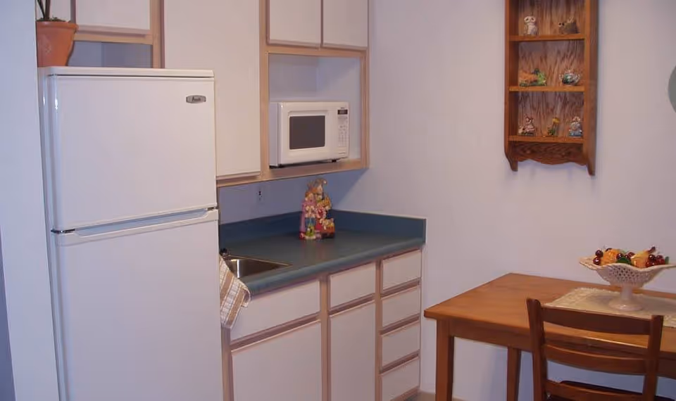 A small kitchen area with a white refrigerator, white cabinets, a microwave, a sink, and a wooden table with a chair. There is a decorative fruit bowl on the table and a wooden wall shelf with small figurines on the wall.
