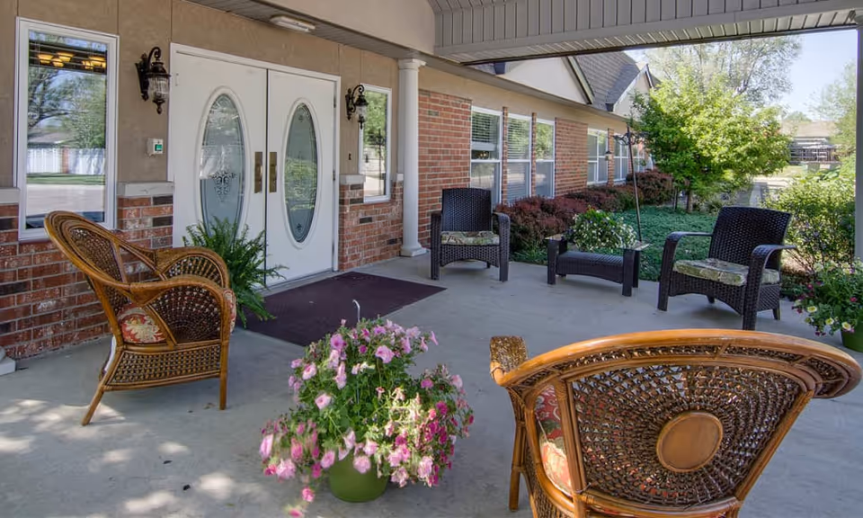 Covered front porch with wicker and rattan chairs, potted flowers, and double decorative glass entrance doors.