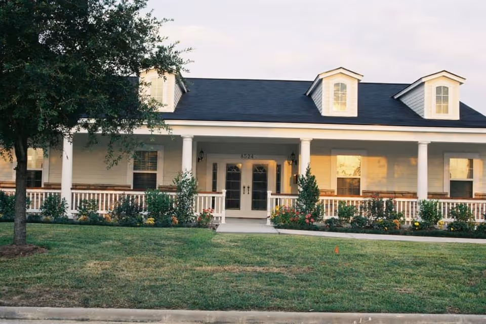 Front exterior view of a single-story building with a dark shingled roof, white siding, and a covered porch supported by white columns. The porch has a white railing and is decorated with various shrubs and flowers. There are three dormer windows on the roof and a tree on the left side of the image. The building is set behind a well-maintained lawn.