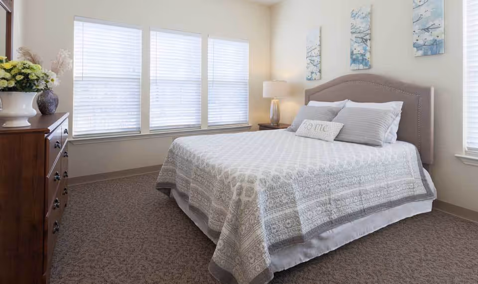 A neatly made bed with a patterned gray and white bedspread and pillows in a bright bedroom. The room has three large windows with blinds, a wooden dresser with a vase of flowers, a bedside table with a lamp, and three decorative wall art pieces above the bed.