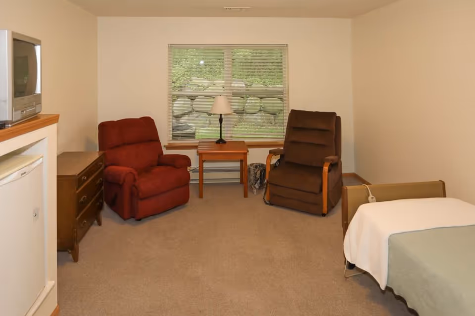 A simple assisted-living room with two recliners, a small table and lamp by a window, a dresser with a TV, and a partially visible bed.