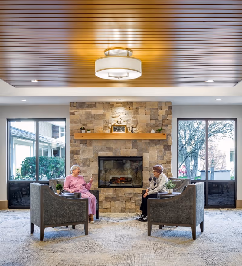 Two elderly women sitting in armchairs facing each other in a cozy living room with a stone fireplace in the center. One woman is wearing a pink outfit and the other is holding a small dog. Large windows on either side of the fireplace let in natural light and show outdoor greenery. The ceiling has wooden paneling with a modern light fixture.