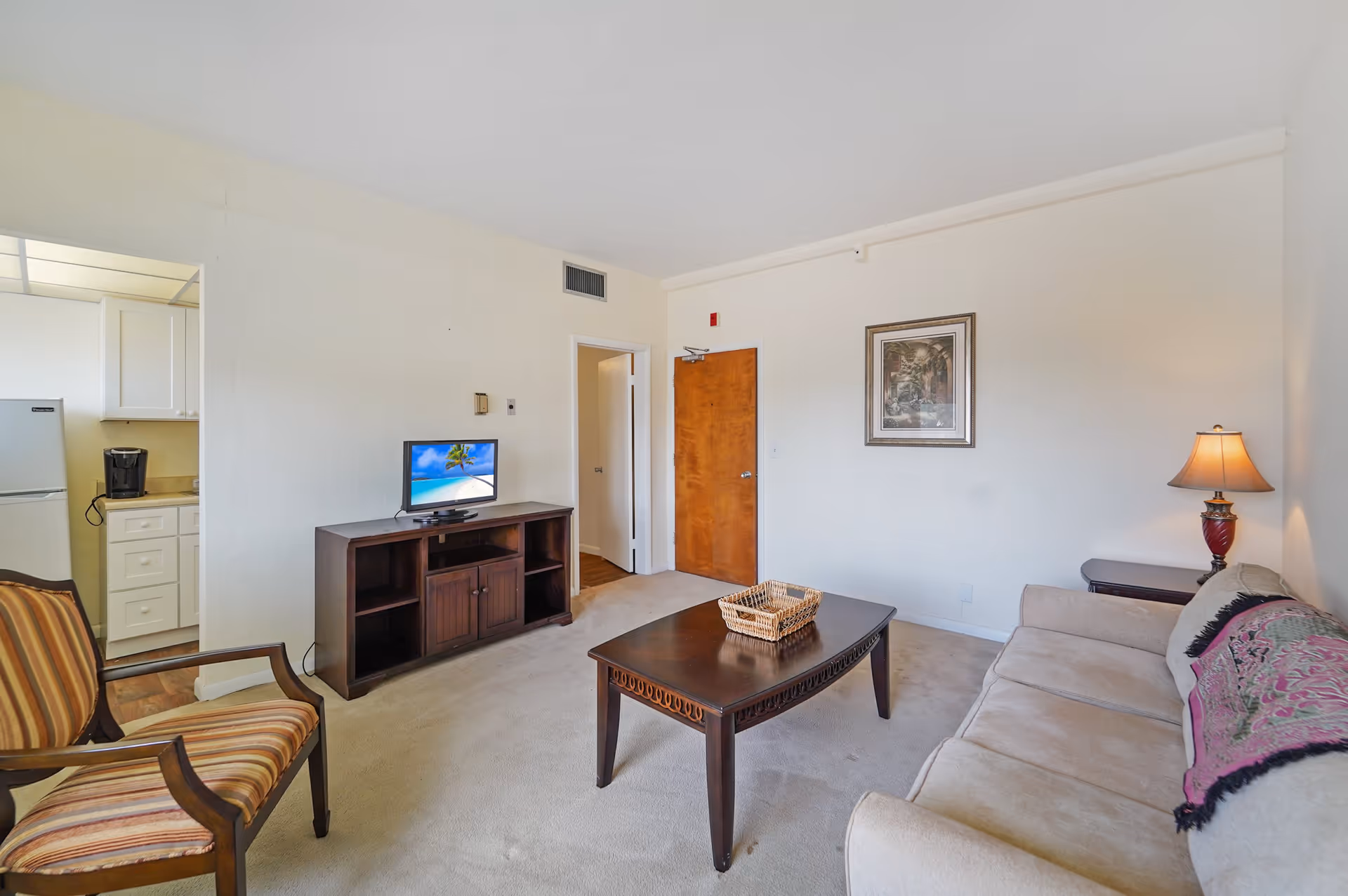 A cozy living room in an assisted living residence featuring a beige sofa with a decorative throw, a wooden coffee table with a woven basket, a striped armchair, a wooden TV stand with a flat-screen TV displaying a beach scene, a side table with a lamp, and a view into a small kitchen area with a refrigerator and coffee maker.
