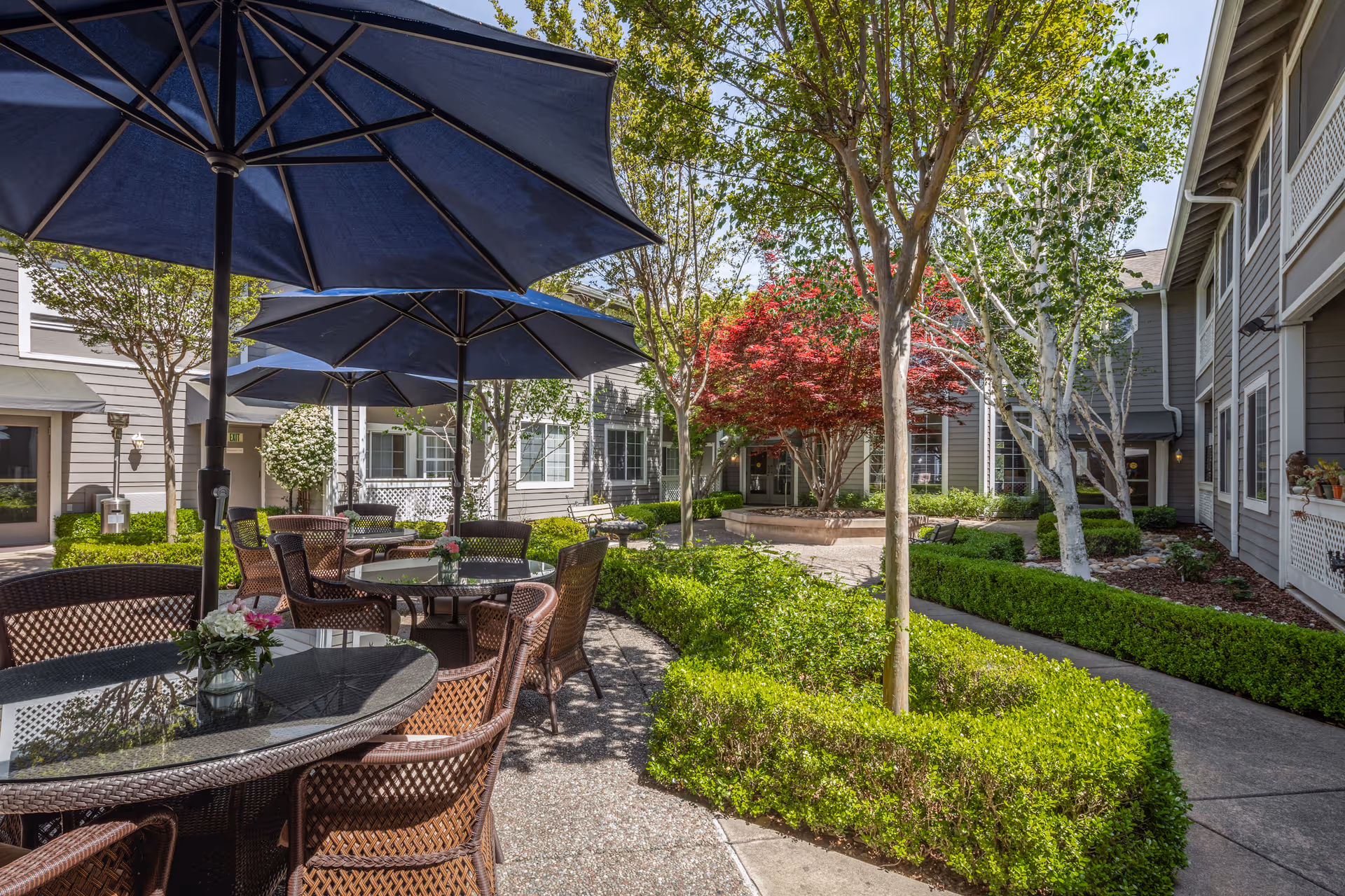 Outdoor courtyard area with round glass-top tables and wicker chairs under large blue umbrellas. The courtyard is surrounded by well-maintained greenery, bushes, and trees, including a vibrant red-leafed tree. The building exterior is visible with gray siding and white trim windows.