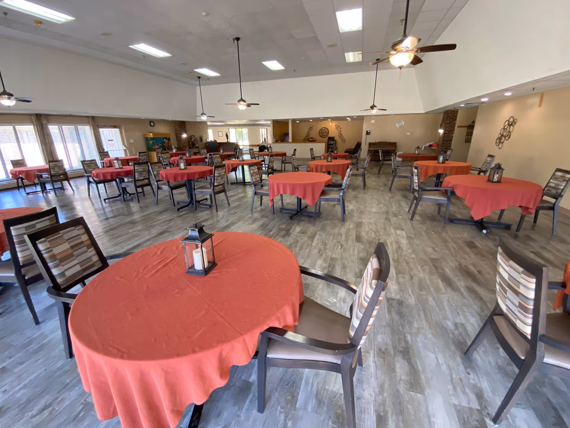 A spacious dining room with multiple round tables covered with red tablecloths, each table surrounded by chairs with striped cushions. The room has a light-colored wood floor, ceiling fans with lights, and large windows letting in natural light. There are decorative wall hangings and a piano against the far wall.