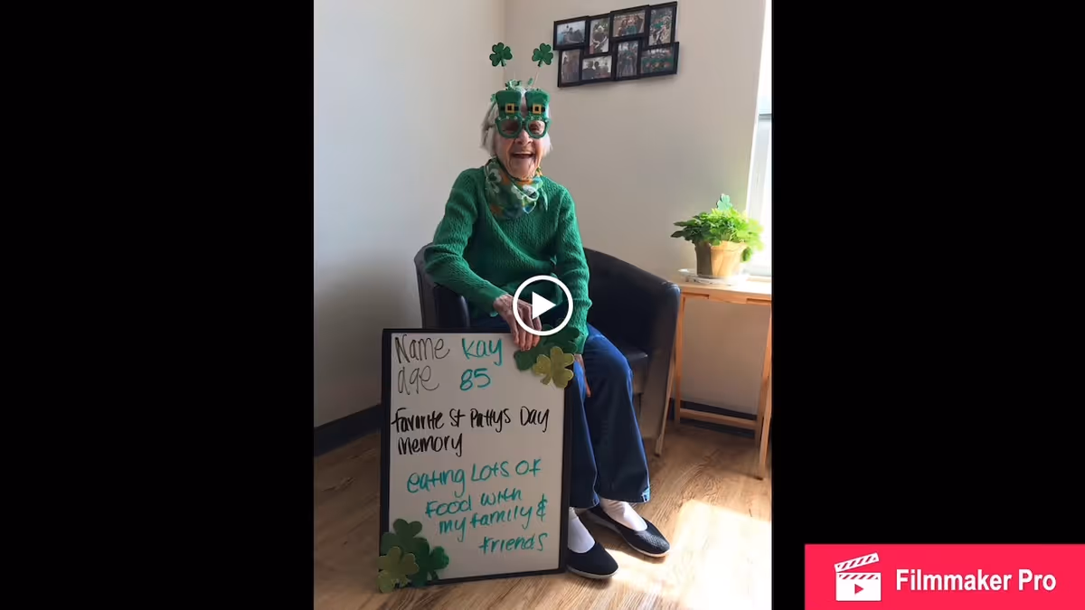 An elderly woman wearing green St. Patrick's Day themed glasses and a green sweater sits in a chair holding a whiteboard. The whiteboard reads: 'Name Kay, age 85, favorite St. Patty's Day memory eating lots of food with my family & friends.' There is a small table with a potted plant next to her and framed photos on the wall behind her.