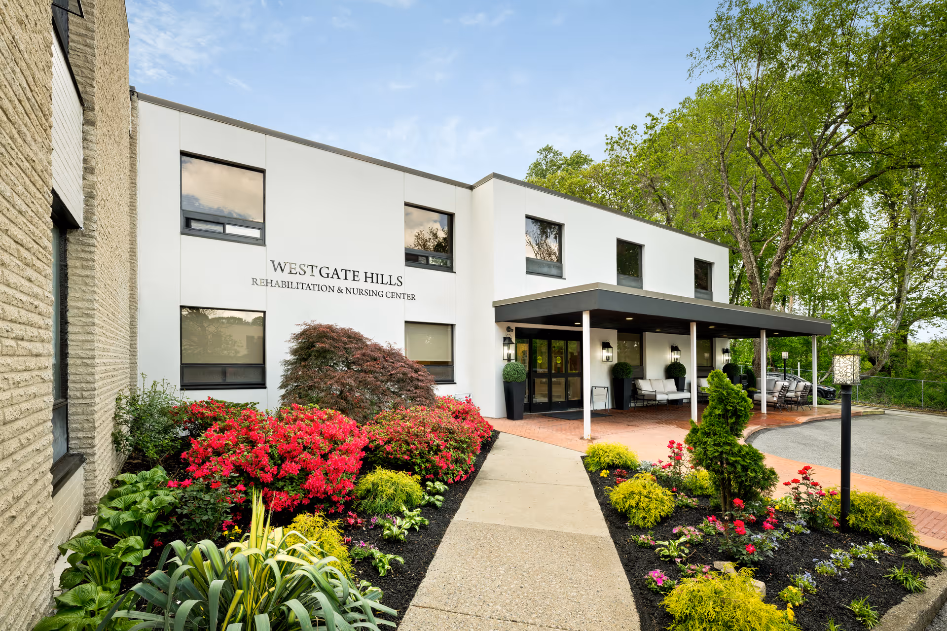 Exterior view of Westgate Hills Rehabilitation & Nursing Center showing a modern two-story building with large windows, a covered entrance with seating, and well-maintained landscaping featuring colorful flowers and green shrubs.
