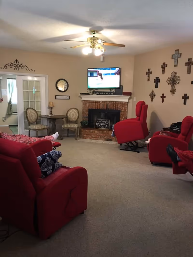 A cozy senior living room with beige walls and carpeted floor. The room features a brick fireplace with a TV mounted above it, displaying a sports game. There are three red recliner chairs, one with a person’s legs visible, and a small table with two upholstered chairs near a glass-paneled door. The wall to the right is decorated with a collection of crosses. A ceiling fan with lights is mounted on the ceiling.