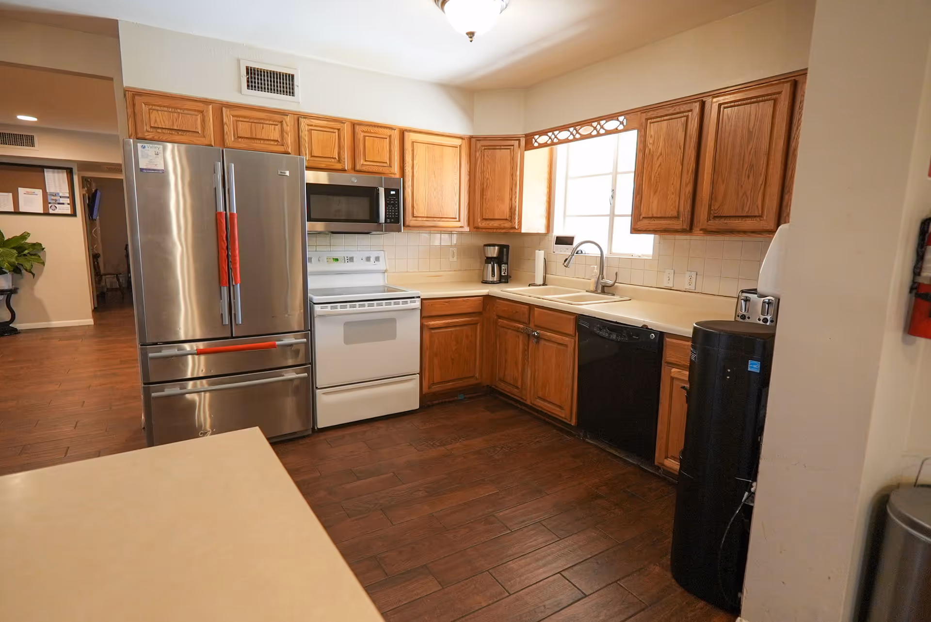 A kitchen area with wooden cabinets, a stainless steel refrigerator, a white stove with an oven, a microwave above the stove, a coffee maker on the counter, a double sink under a window, a dishwasher, and a black appliance on the floor. The floor is dark wood, and the walls are light-colored.