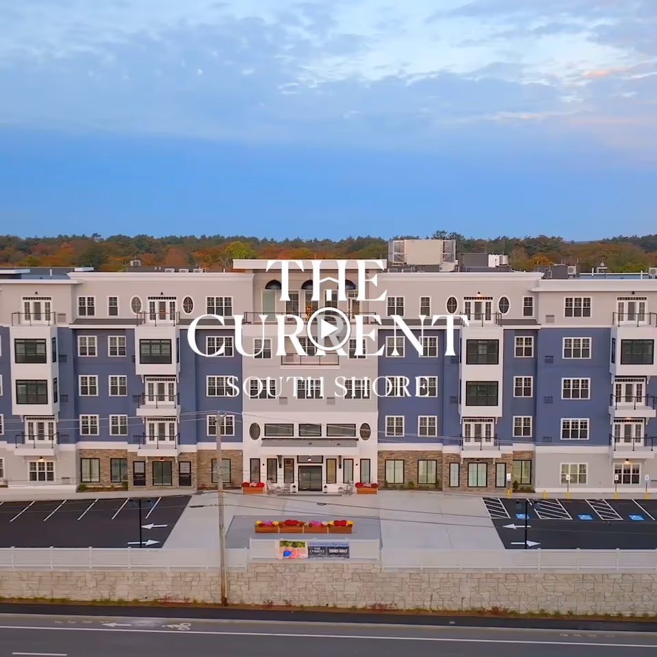 Exterior view of The Current South Shore, a multi-story senior living facility with a modern design featuring blue and white walls, multiple windows, balconies, and a parking lot in front. Trees and a partly cloudy sky are visible in the background.