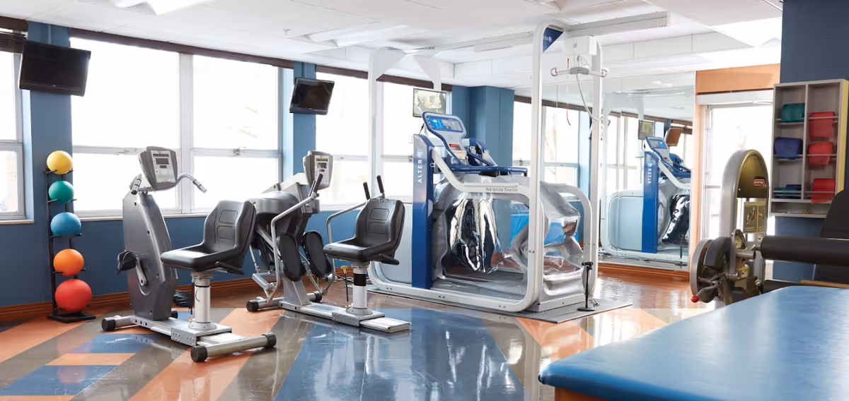 A bright exercise room with large windows, featuring various fitness equipment including two recumbent exercise bikes, an anti-gravity treadmill, a weight machine, and a rack of colorful exercise balls. The room has a polished floor with a geometric pattern and mirrors on one wall.