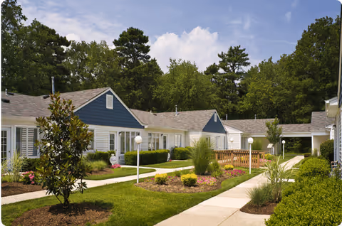 Outdoor view of a senior living facility with single-story buildings featuring blue and white siding, surrounded by well-maintained landscaping including green lawns, bushes, flowers, and trees under a partly cloudy sky.