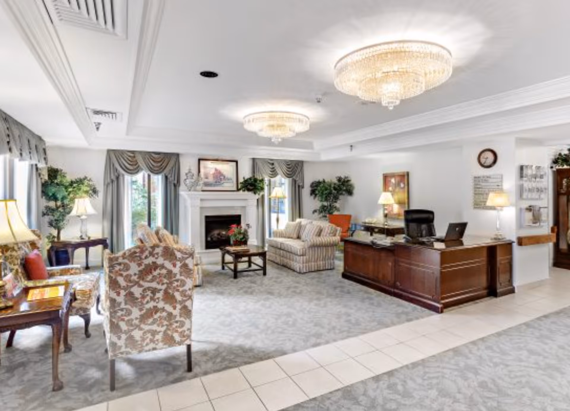 Spacious, well-lit senior living common area with seating, a fireplace, chandeliers, and a reception desk.