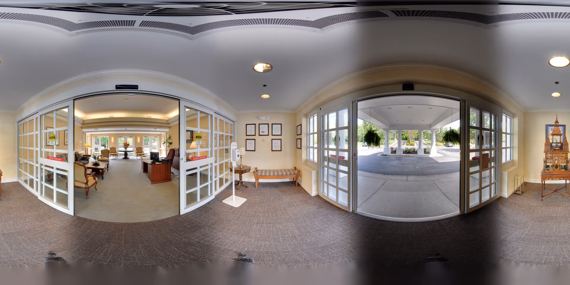 Entrance area of a senior living facility showing automatic sliding glass doors leading outside to a covered driveway with hanging plants. Inside, there is a reception area with a desk, chairs, and a bench along the wall. The space is well-lit with ceiling lights and has framed certificates on the wall.