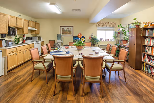 A well-lit dining room with a large rectangular table surrounded by chairs. The room features wooden flooring, a kitchenette area with cabinets, microwave, and coffee maker on the left side. There are plants and decorative items on the table and shelves, a water cooler, and a window with a valance letting in natural light.