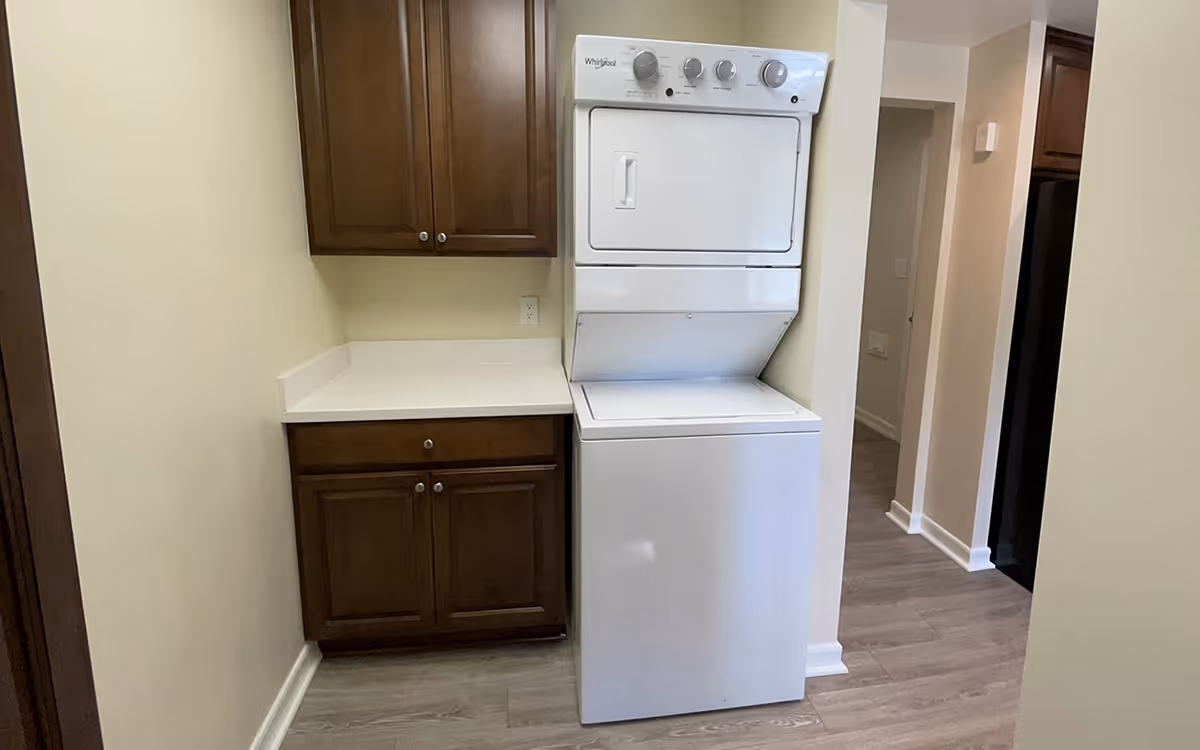 Laundry area with a white stacked Whirlpool washer and dryer unit next to brown wooden cabinets with a white countertop. The floor is light wood, and there is a hallway visible to the right.