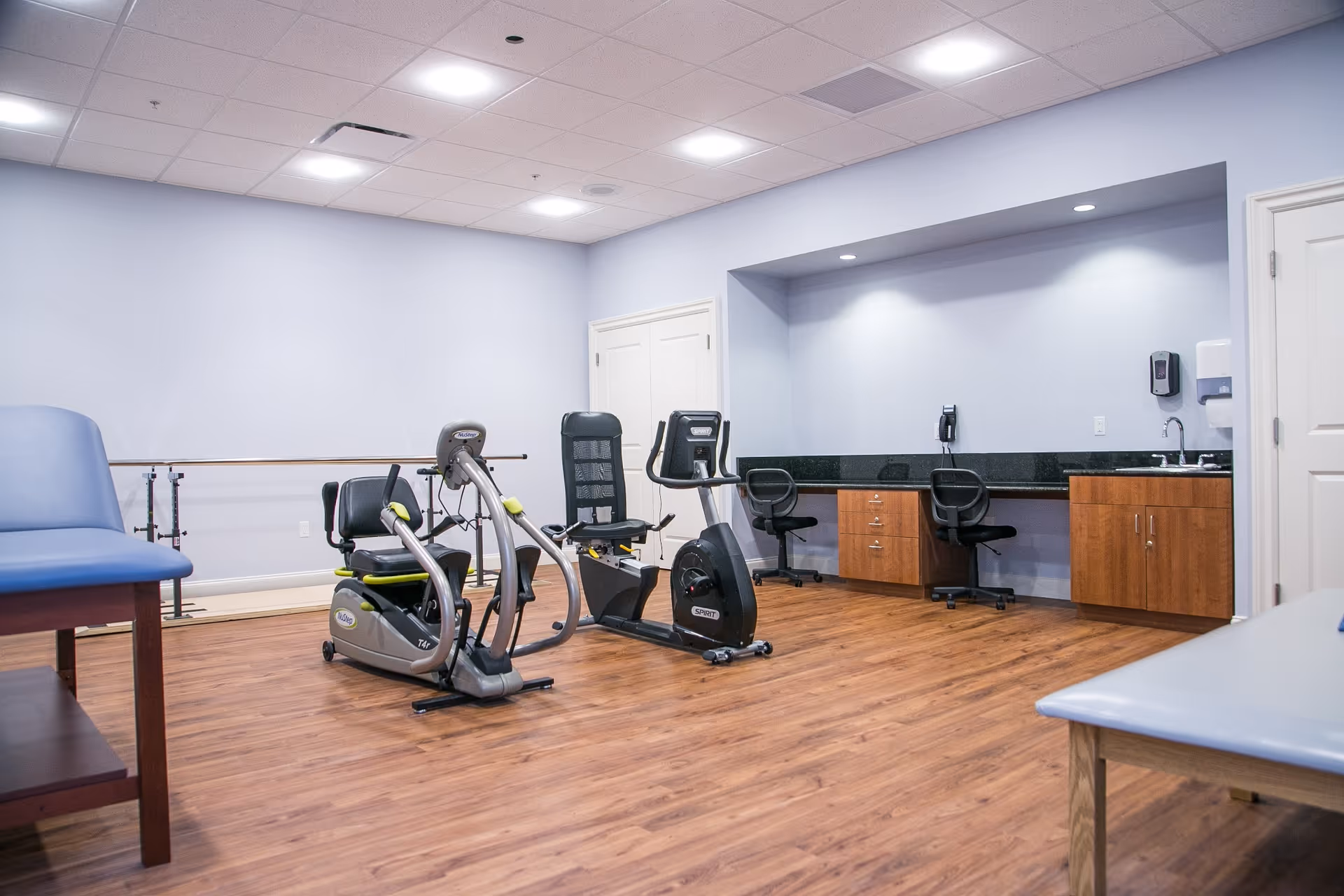 Spacious interior therapy/fitness room with recumbent exercise bikes, treatment tables, wood floors, and a countertop with chairs and cabinets.