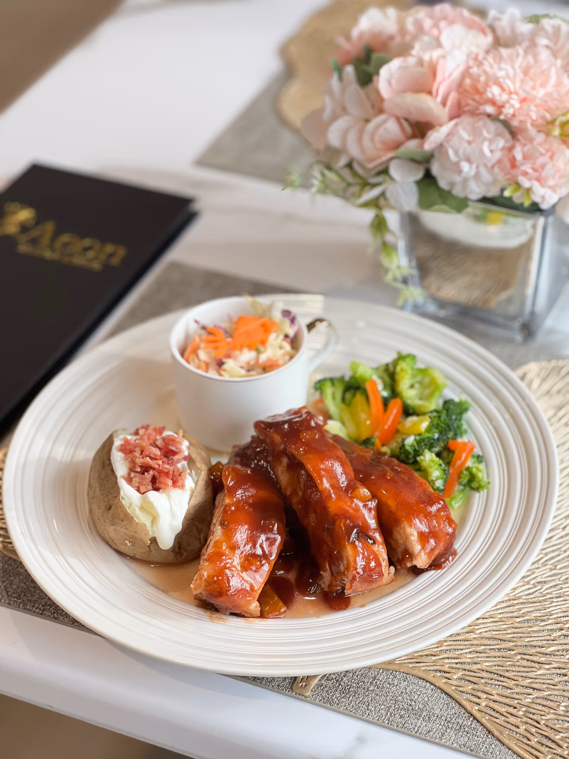 Plate of glazed ribs with a baked potato, mixed vegetables and coleslaw on a dining table with a floral centerpiece.