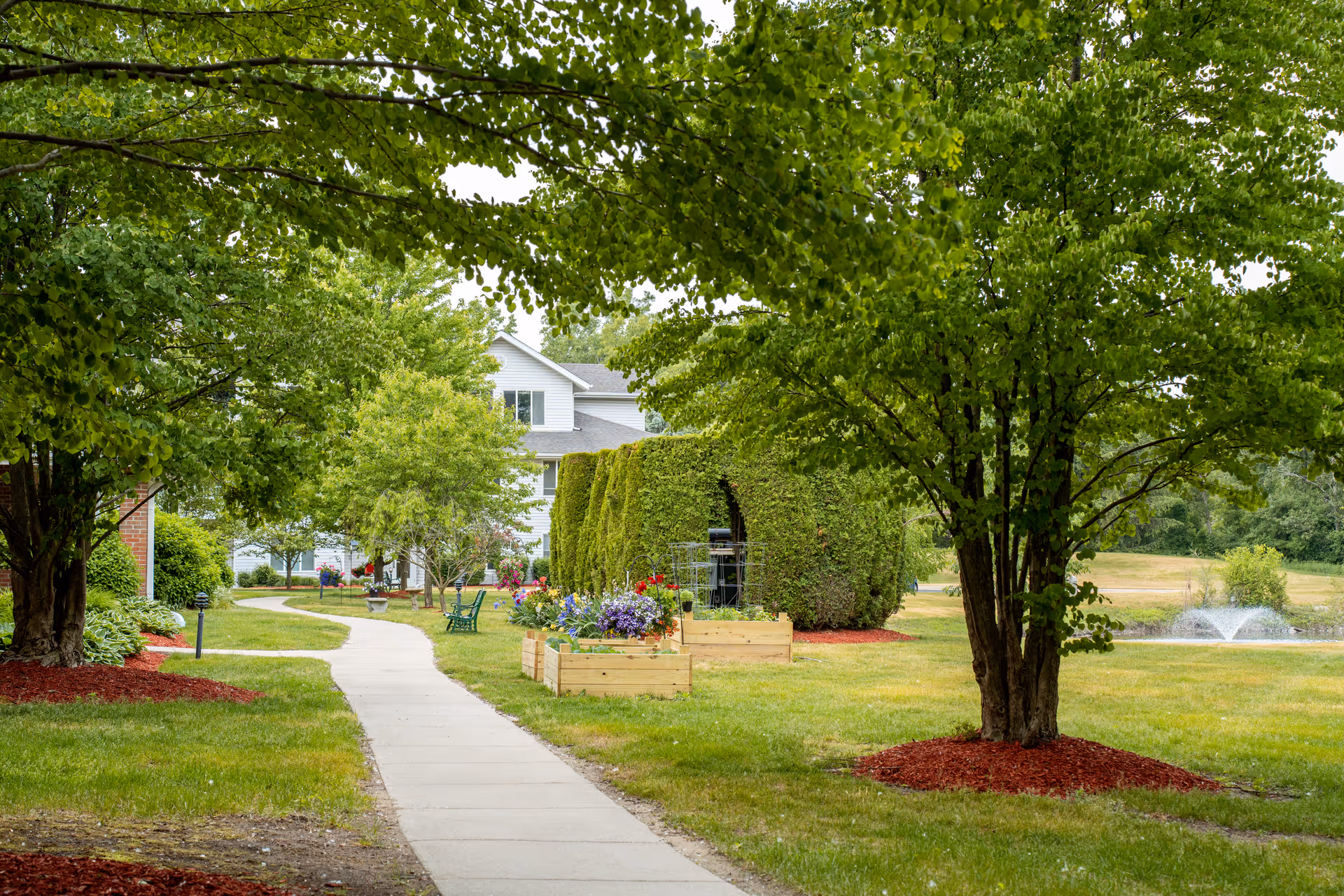 A peaceful outdoor garden area with a paved walkway winding through green grass, trees, and flower beds. There are wooden raised garden boxes with colorful flowers, neatly trimmed hedges, and a water fountain in the background. A white building is partially visible behind the trees and hedges.