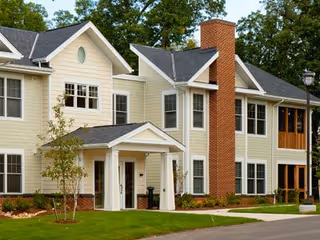 Exterior view of a multi-story residential building with beige siding, white trim, and a prominent red brick chimney. The building has multiple windows, a small covered entrance with white columns, and is surrounded by green grass and trees.