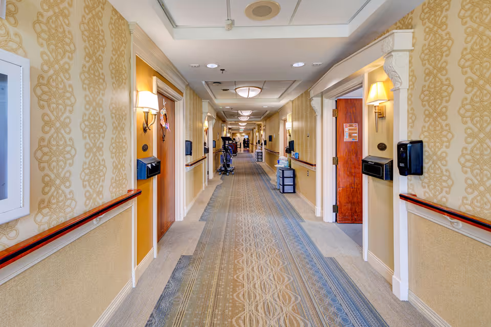 A long, well-lit hallway in a senior living facility with patterned wallpaper and carpet. The hallway features wooden doors on both sides, each with a room number and a wall-mounted light above. Handrails run along the walls, and there are various medical and cleaning supplies placed along the corridor. The ceiling has recessed lighting and ventilation fixtures.