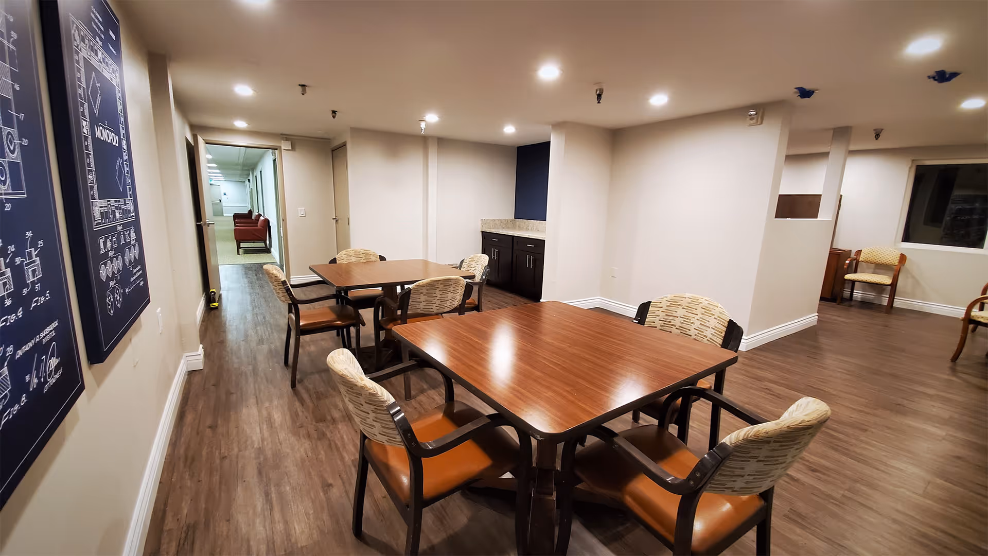 Interior view of a senior living facility common area with wooden tables and cushioned chairs arranged for seating. The room has wood flooring, recessed ceiling lights, and light-colored walls. There are framed blueprints or diagrams on one wall and a hallway leading to other rooms in the background.