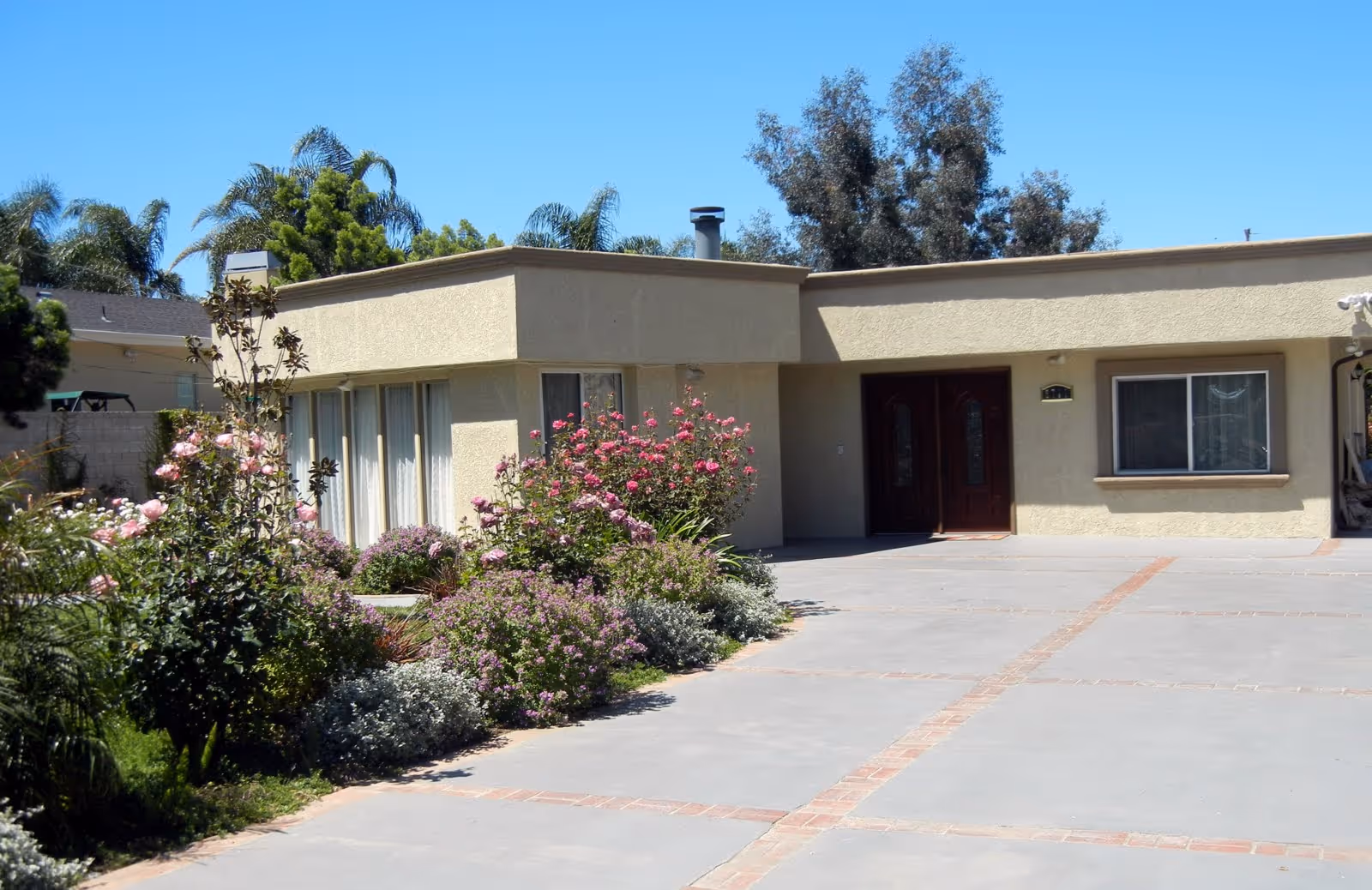 Single-story beige building with double front doors, a wide driveway, and flowering shrubs in the front yard.