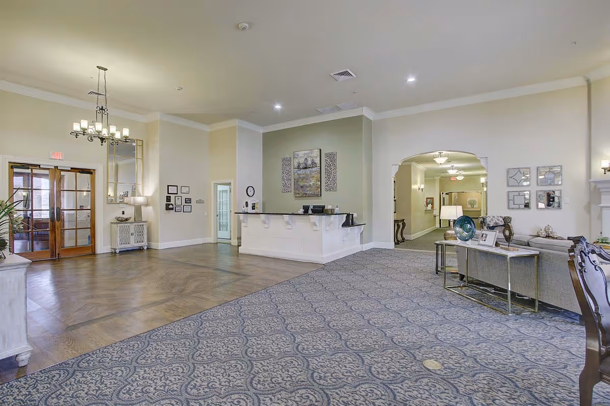 Spacious and well-lit senior living facility lobby with a reception desk, patterned carpet, wooden flooring near the entrance, chandelier lighting, and comfortable seating area with a sofa and decorative items.