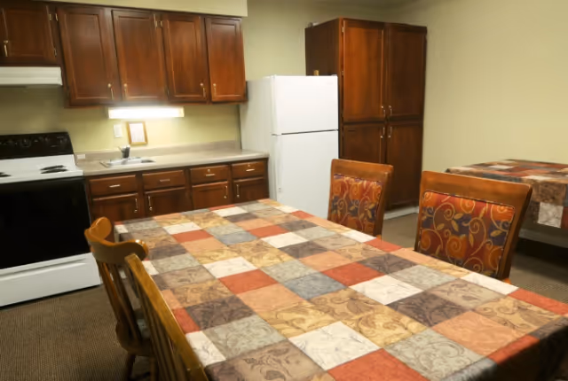 Interior view of a kitchen and dining area with a table covered by a multicolored patchwork tablecloth, surrounded by wooden chairs with patterned cushions. The kitchen has wooden cabinets, a white refrigerator, a white stove, and a countertop with a sink.