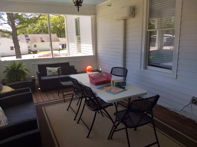 A screened-in porch area with a white folding table surrounded by six black chairs. On the table are a pink plastic container, a box of tissues, and some coloring books with crayons. There are two black cushioned sofas with pillows along the walls, a potted fern, and a hanging light fixture on the ceiling. The porch has white walls, wooden flooring, and large windows showing an outdoor view with trees and buildings.