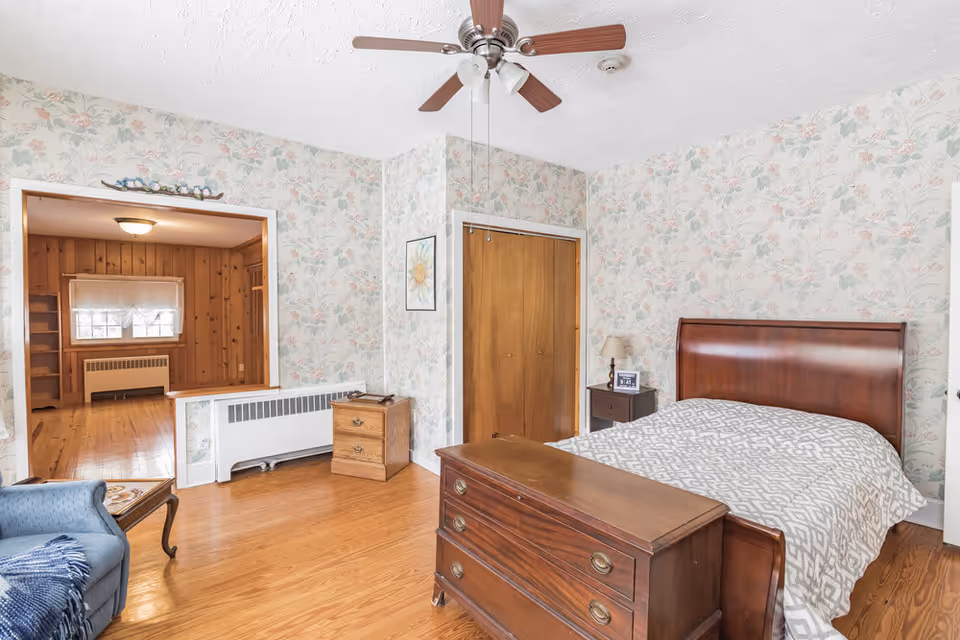 A bedroom with floral wallpaper, a wooden bed with a patterned bedspread, a wooden chest at the foot of the bed, a nightstand with a lamp and a framed photo, a wooden closet door, and a ceiling fan with wooden blades. There is an open doorway leading to another room with wood-paneled walls and a window with a white curtain.
