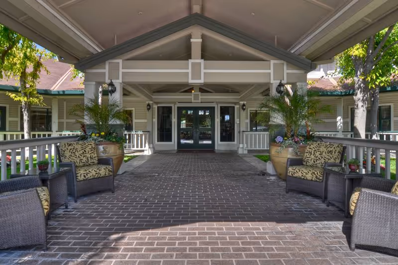 Covered entrance area of a senior living facility with a brick pathway, wicker chairs with patterned cushions, large potted plants, and double glass doors leading inside. The building exterior is light-colored with green trim and surrounded by trees and landscaping.