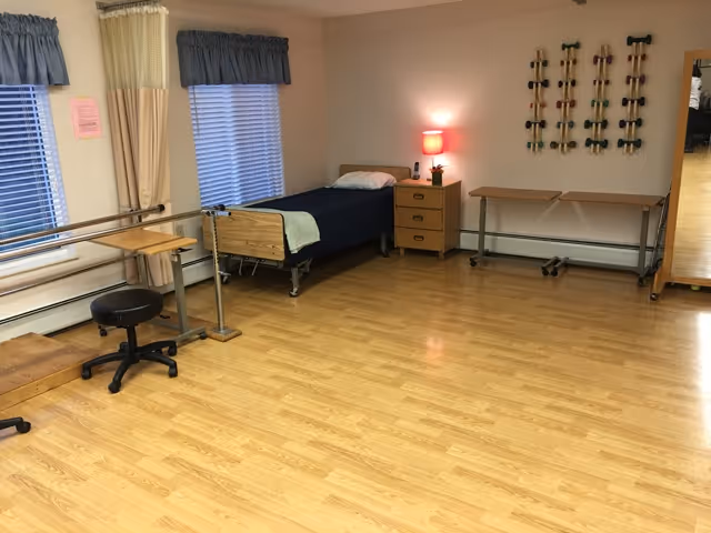 A room with light wood flooring featuring a single hospital-style bed with a blue blanket and pillow, a small wooden nightstand with a lamp and a plant, two windows with blue valances, a set of parallel bars for physical therapy, a black rolling stool, a table, and a wall-mounted rack holding multiple pairs of hand weights.