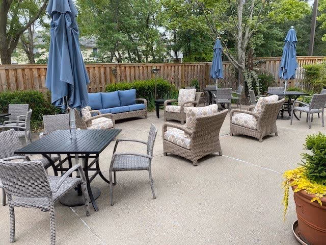 Outdoor patio area with multiple wicker chairs and tables, some with blue umbrellas. There is a blue cushioned sofa and several armchairs arranged for seating. The area is surrounded by a wooden fence and trees, with a large potted plant with yellow flowers in the foreground.