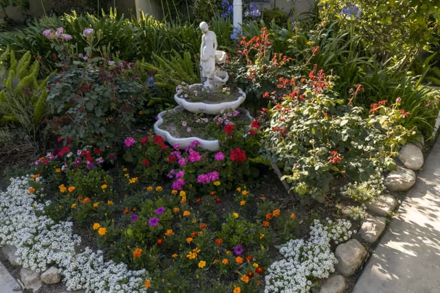 A colorful garden bed with various flowers including white, orange, purple, pink, and red blooms surrounding a tiered stone fountain topped with a statue of a woman. The garden is bordered by stones and is adjacent to a concrete pathway.