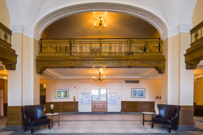 Spacious historic lobby with a mezzanine balcony, chandeliers, and two leather armchairs flanking a small table.